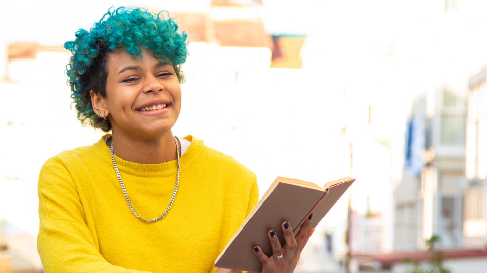 Hispanic high schooler with blue curly hair seated, reading a book and smiling off camera.