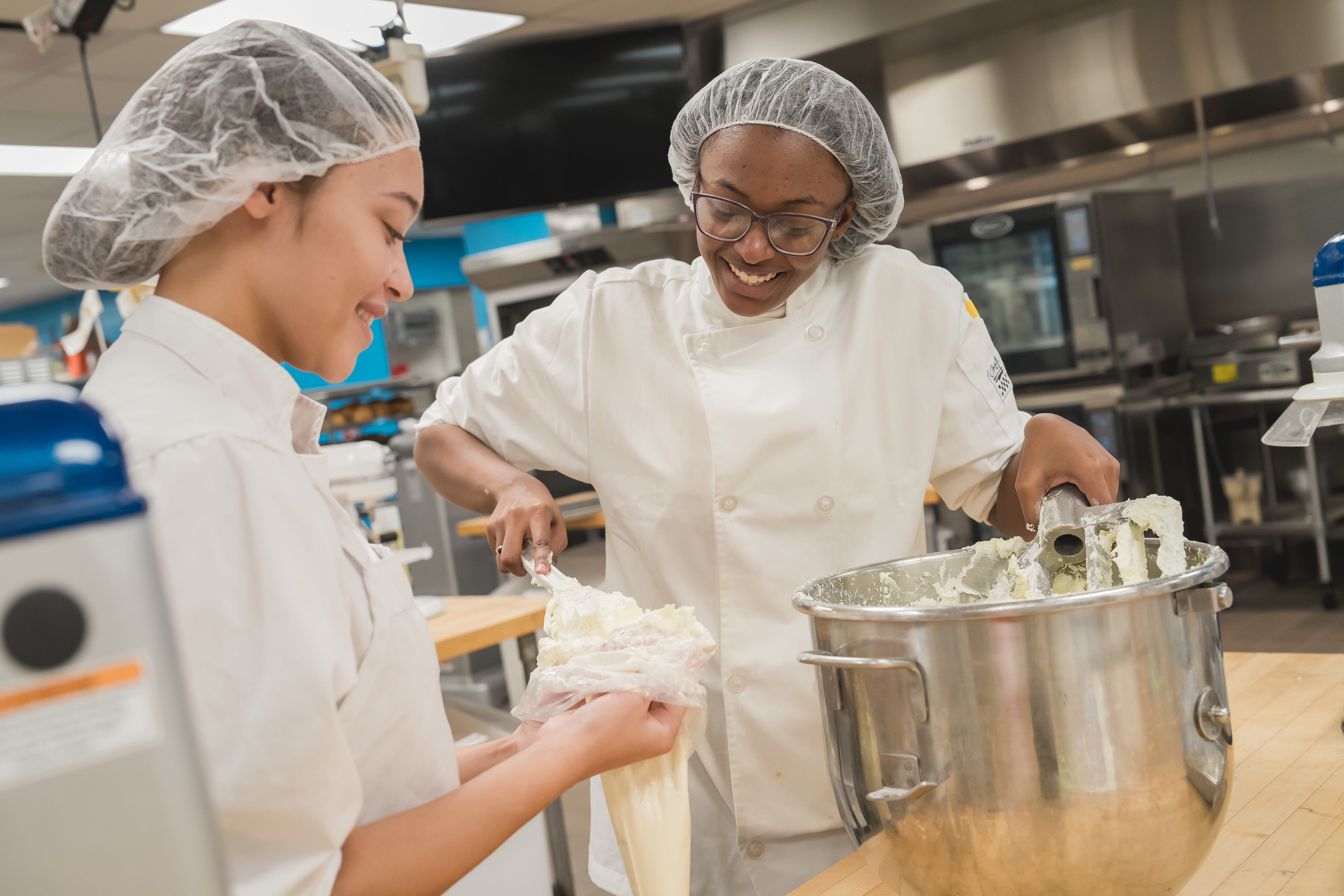 Two CTE culinary students from Genesee County Technical Campus are standing at a table in a professional kitchen. They are both wearing white chef uniforms and are mixing contents to put in a piping bag.
