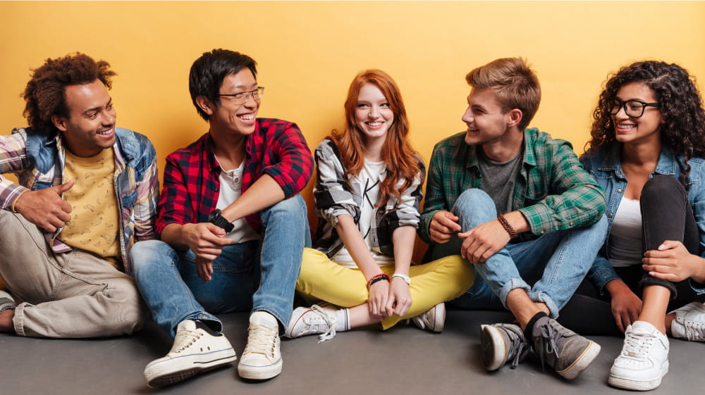 Diverse group of high schoolers seated against a wall, smiling and talking.