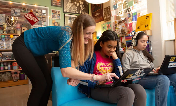 two middle school girls sitting on couch working on computers as teacher leans over on pointing to the screen