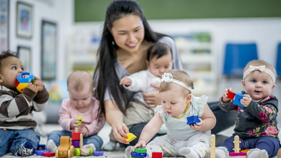 Caregiver holding infant while surrounded by four infants playing with blocks