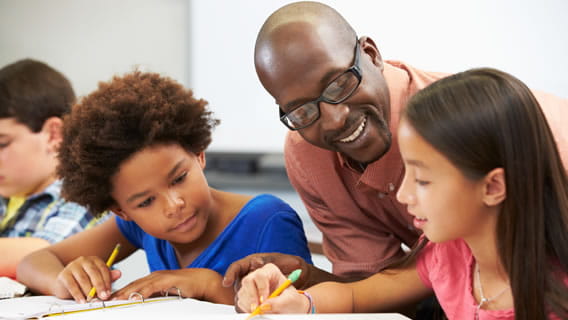 two students doing school work as teacher leans over smiling and encouraging
