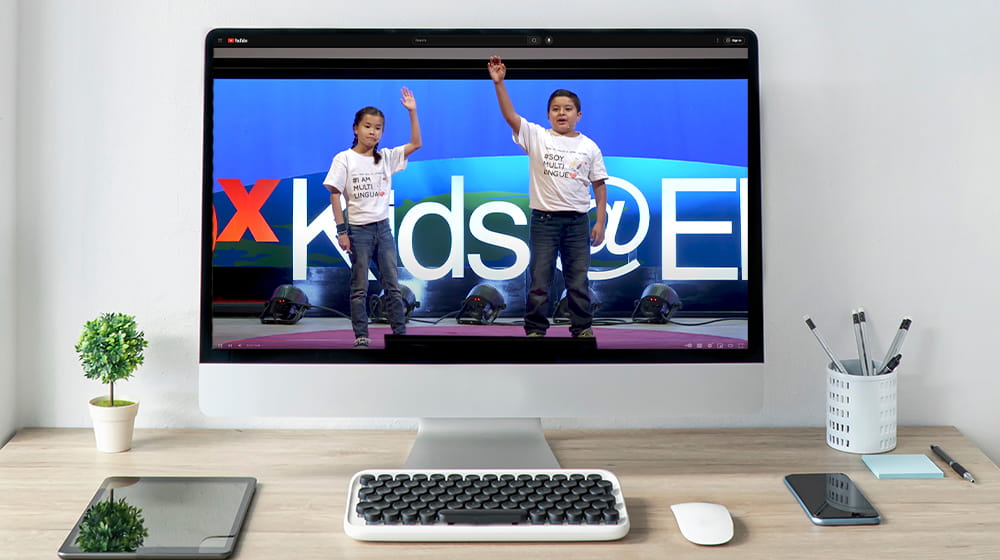 Computer on desk with screen showing young female and male Hispanic students presenting a TED talk.