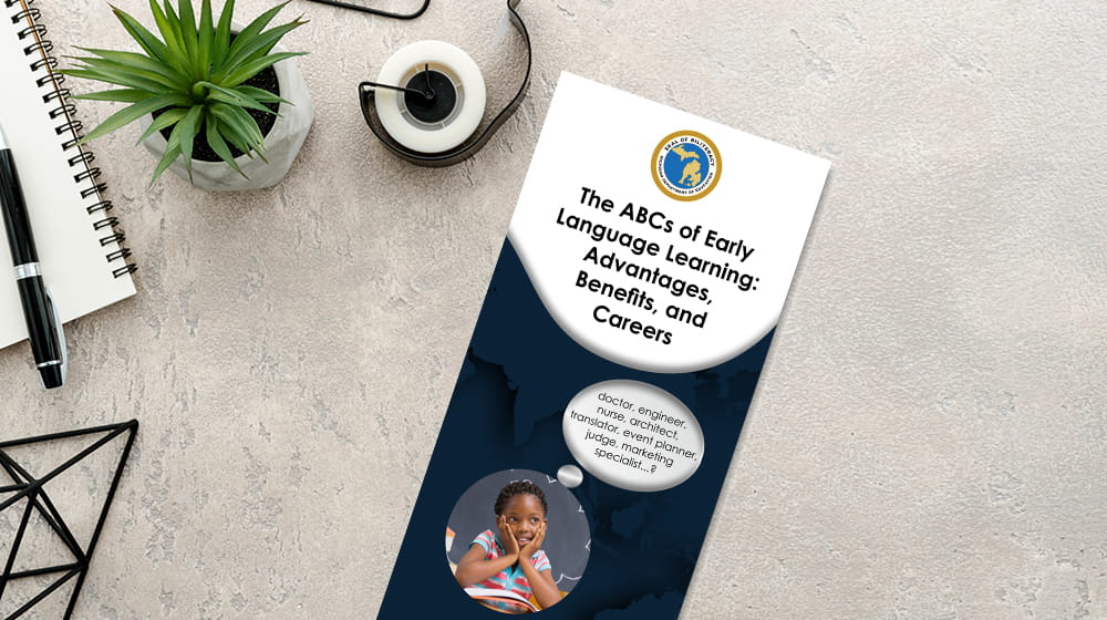 The ABCs of Early Language Learning brochure on light gray desk surrounded by a notepad and pen and tape dispenser.