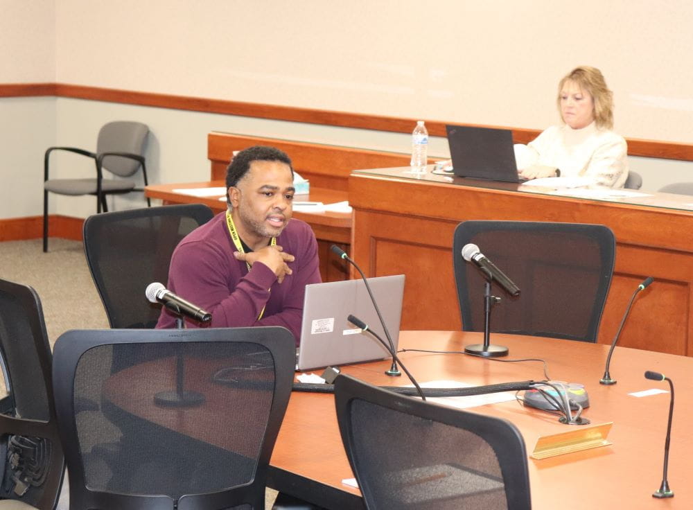 African-American man sitting at end of a board table with lap top and open hand to chest.
