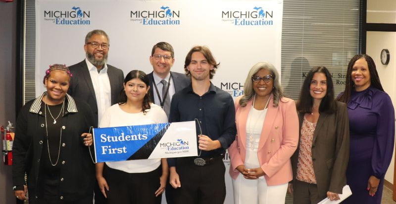 Students and adults holding blue and white 'students first' banner in front of blue and white MDE backdrop