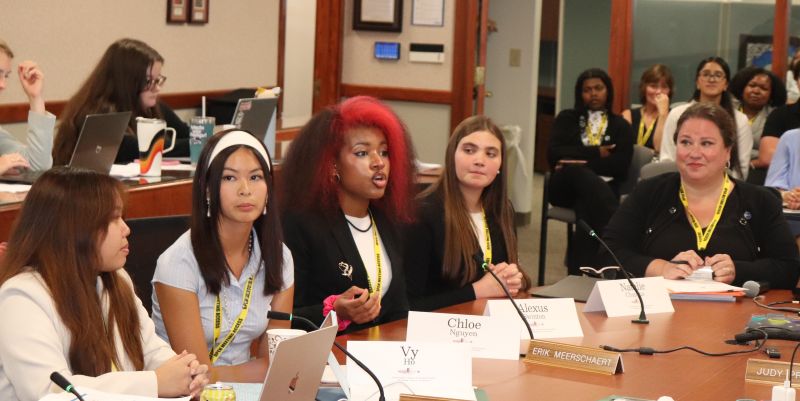 4 students and adult sitting at board table with microphones and table tents in front of them