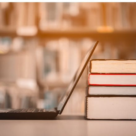 A blurry stack of library shelves in the background, a table in the foreground with a side view of an open laptop next to a stack of four books seen from the bottom