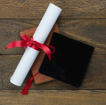 Top-down view of a brown leather book on a wooden table. On top of the book is a white rolled-up graduation certificate tied with a red ribbon and a black graduation cap with a red tassel.