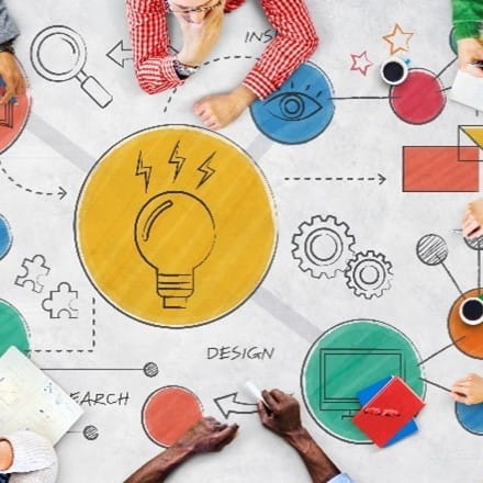Top-down view of several people sitting around a table covered with a large diagram of puzzle pieces, a magnifying glass, and cogs; with images and arrows related to ideas, designs, research, and insight.