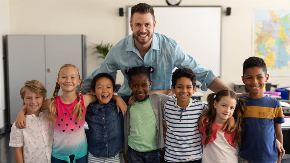 Caucasian male elementary teacher smiling at the camera with a group of diverse young elementary students.