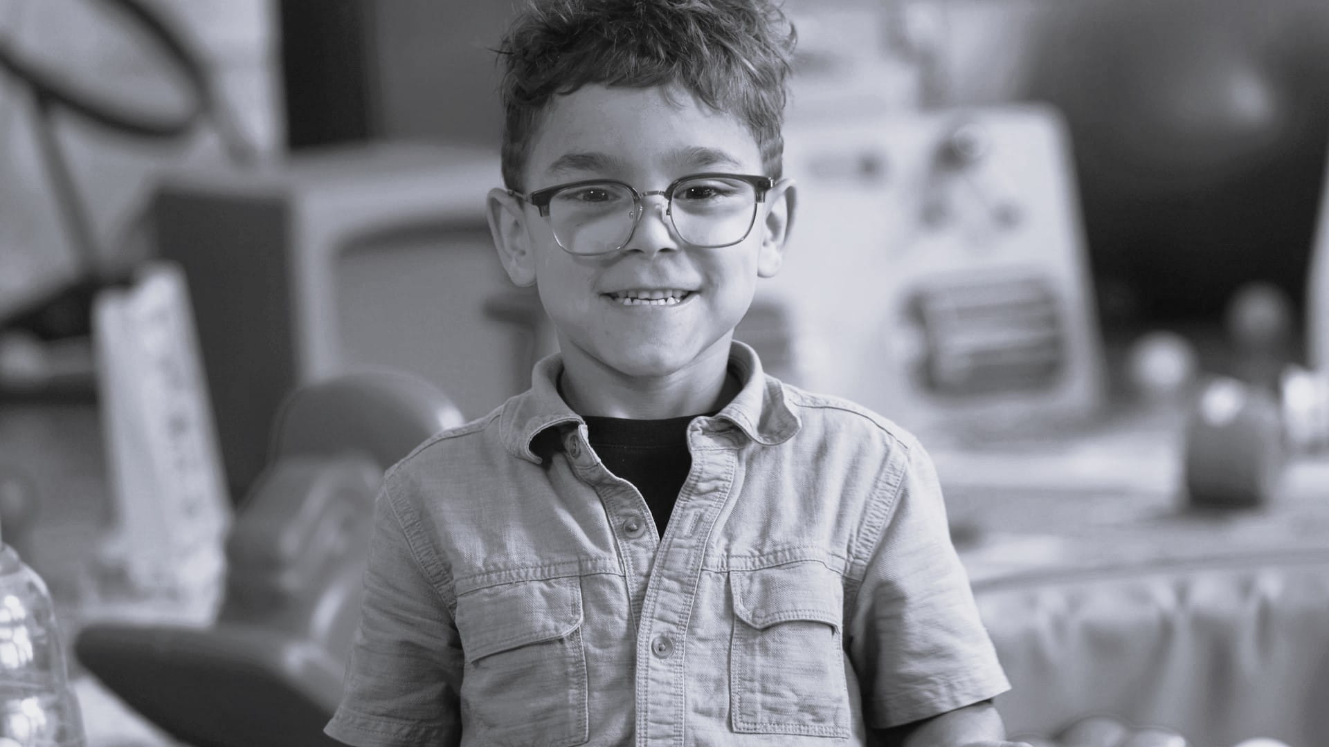 A young boy wearing glasses smiles at the camera while standing in a classroom setting.
