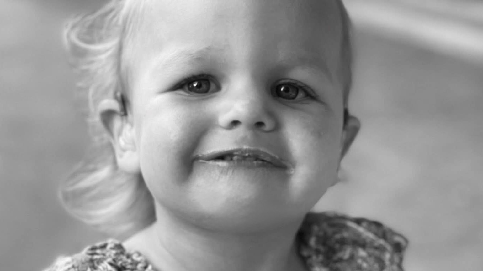 A toddler girl wearing a floral blouse smiles at the camera.