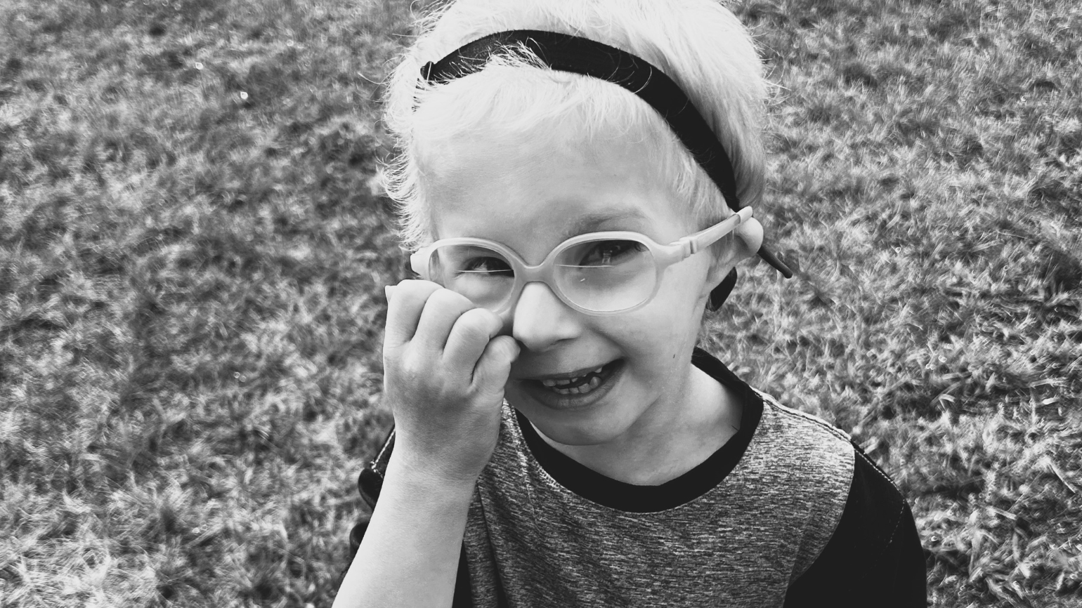 A smiling young boy wearing a bandana and glasses smiles at the camera while holding one hand up to his face.