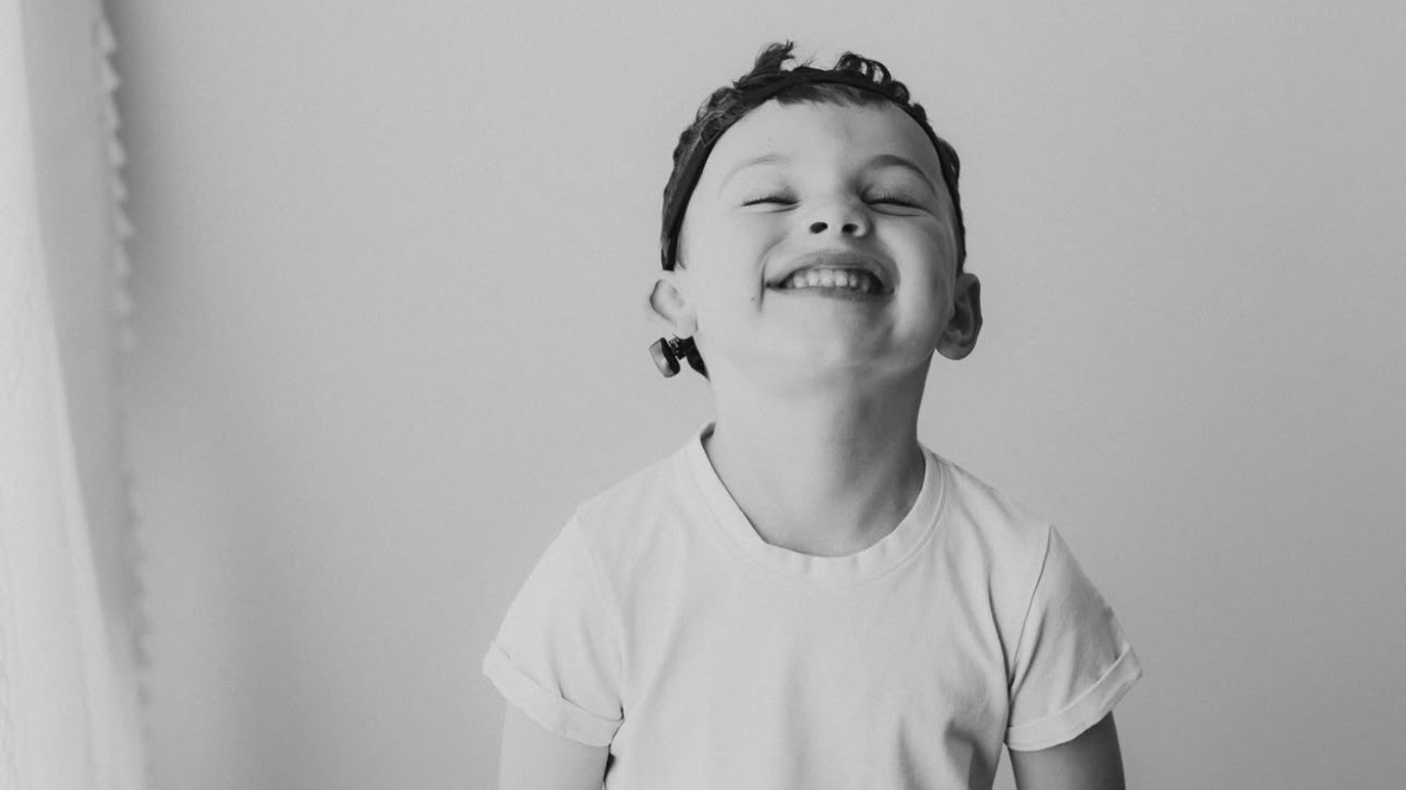 A smiling young boy wearing a white t-shirt smiles while lifting his face upward.