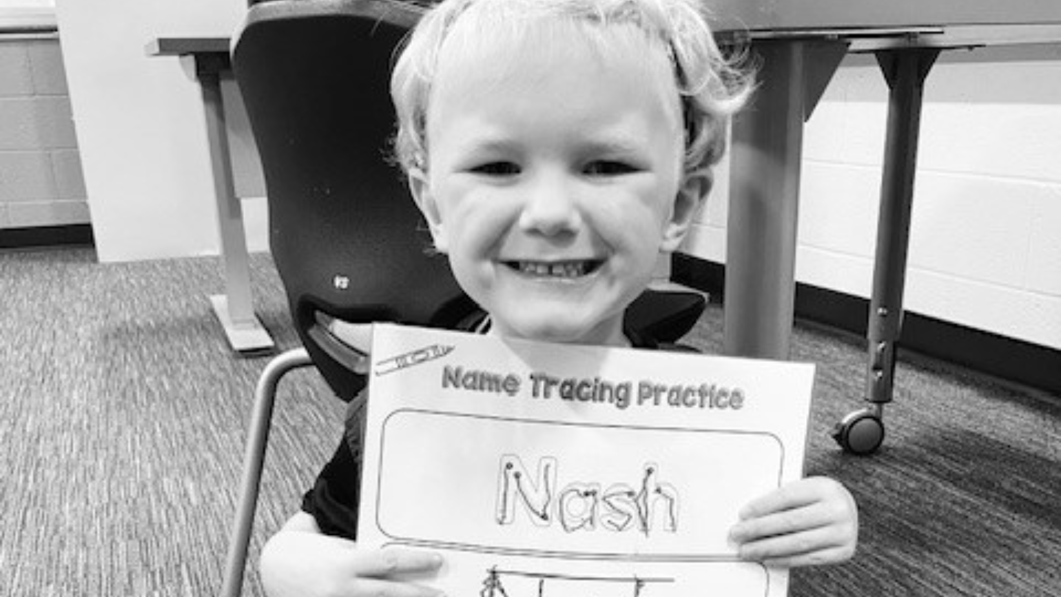 A young boy smiles for the camera while holding a "name tracing practice" sheet with his name, Nash, on it.