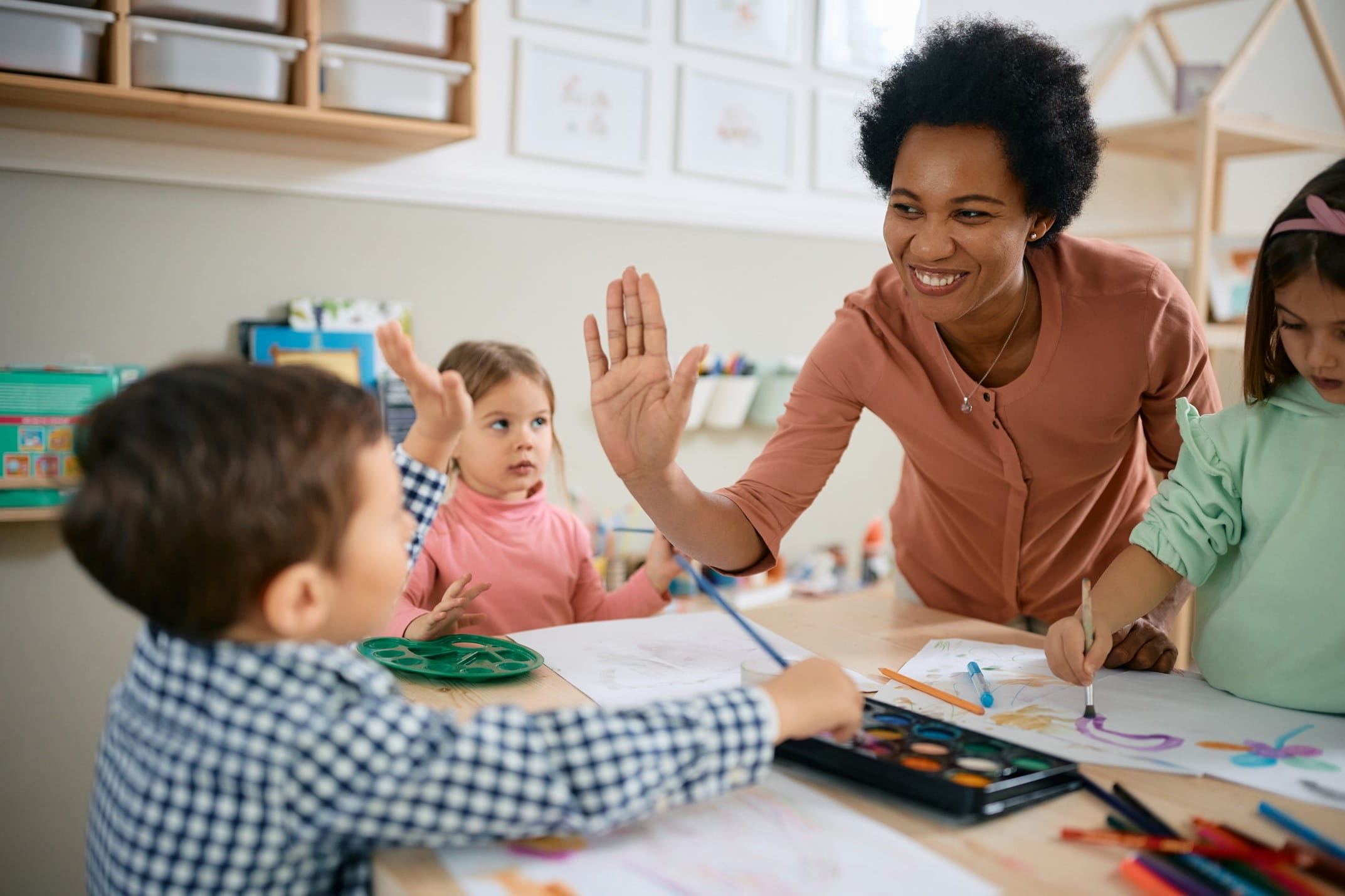 A teacher smiles while high-fiving a young boy doing an art project at a table full of kids in a classroom.