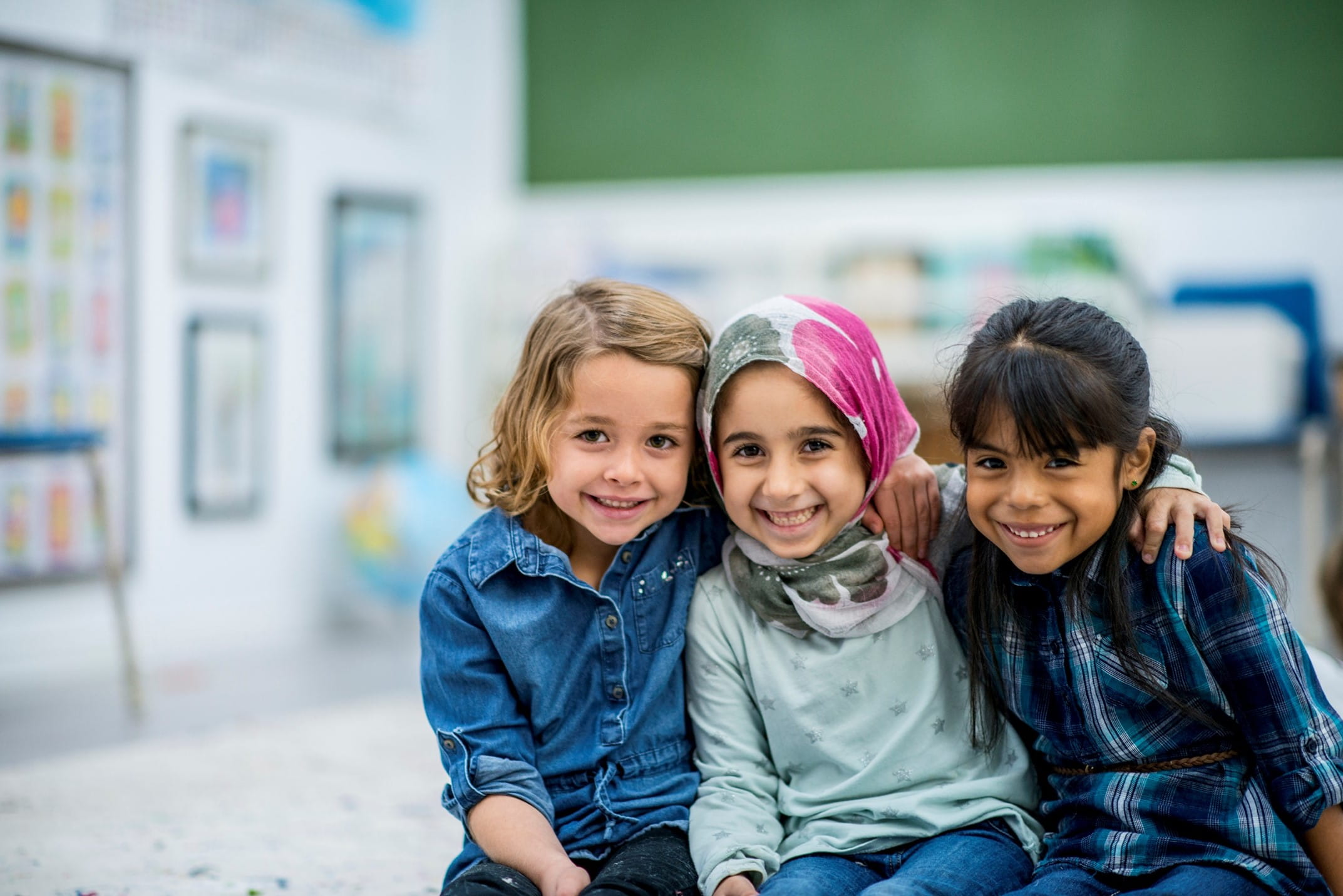 Three young girls in a classroom smile at the camera with their arms around each other's shoulders.