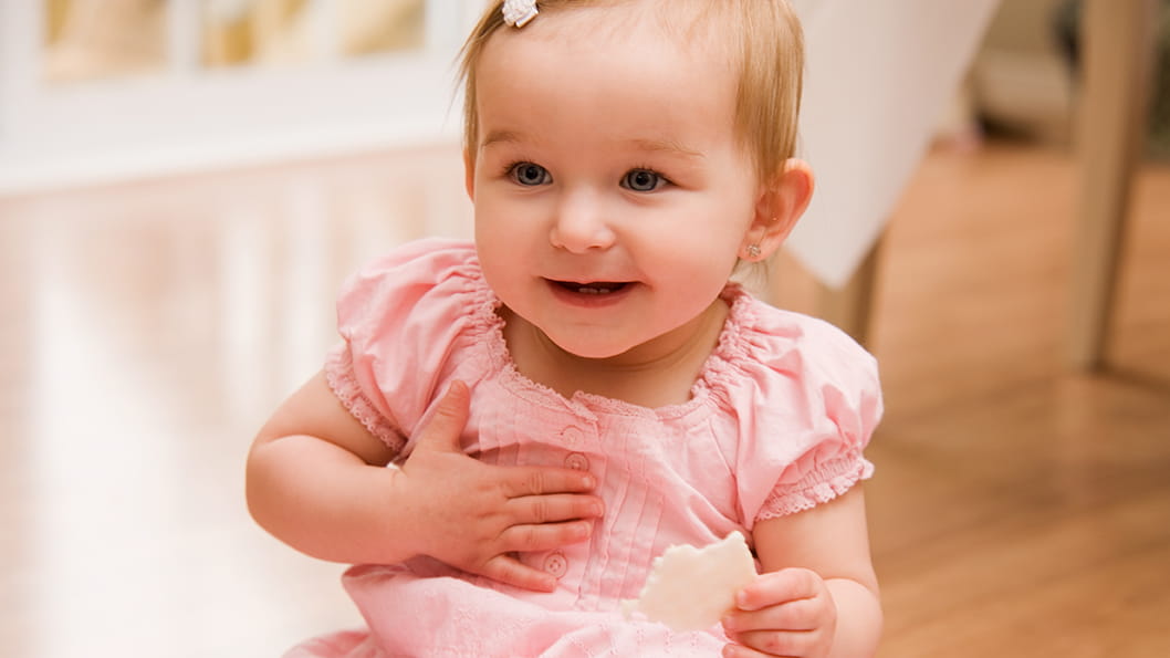 A baby girl wearing a pink dress smiles while holding a hand to her chest.