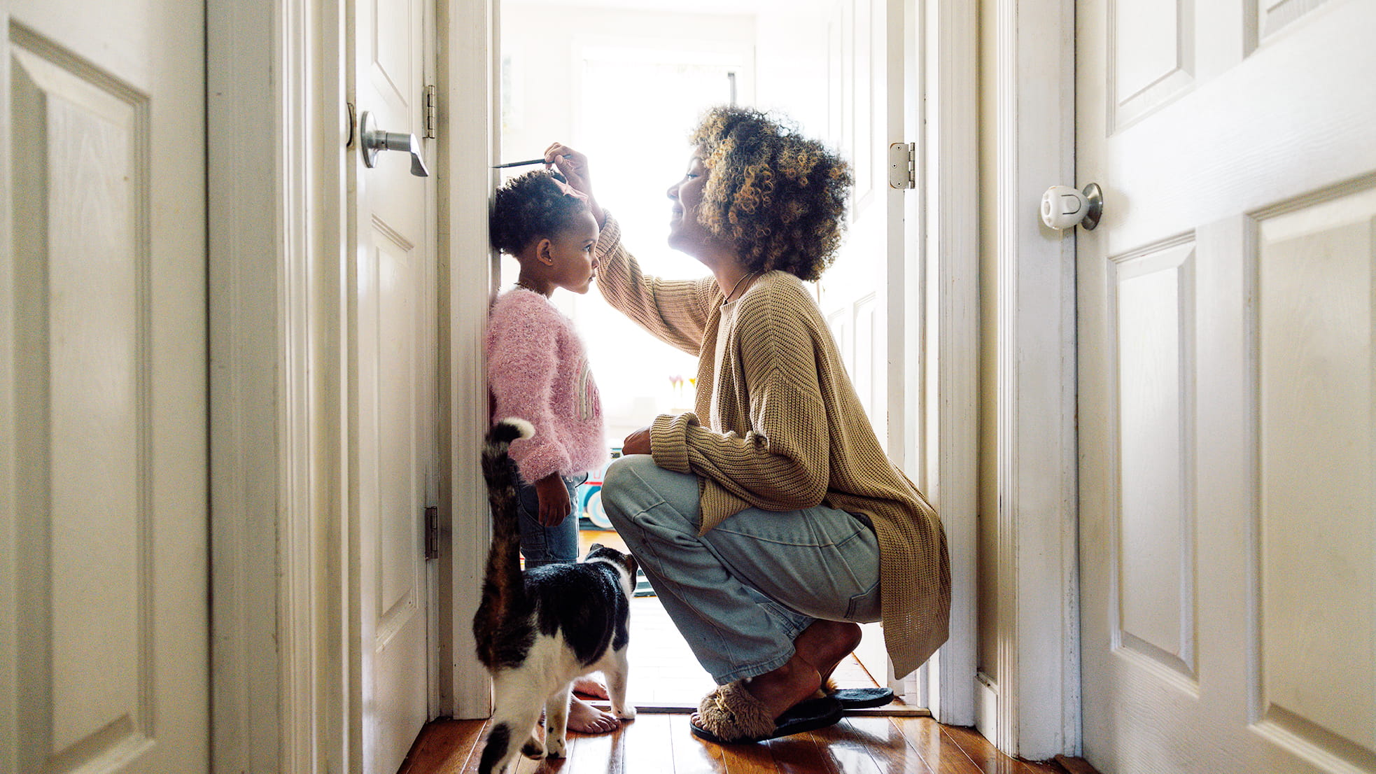 A mother smiles while marking her young daughter's height on a door frame at home as a cat walks past.