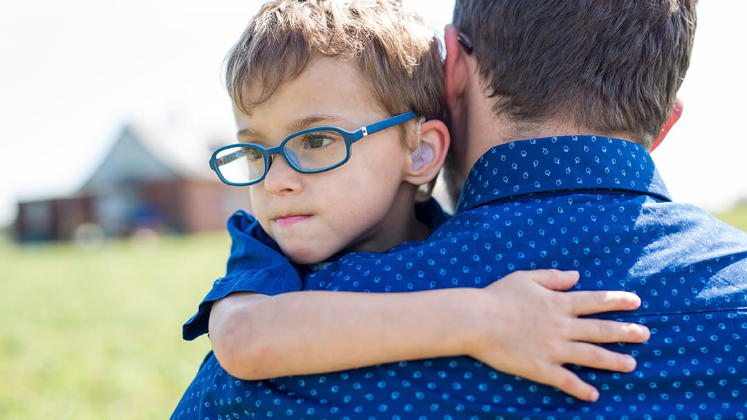 A young boy wearing glasses and a hearing device peers over his father's shoulder while being held.