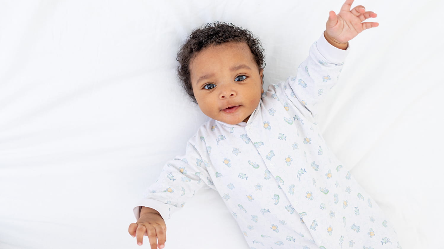 A young baby wearing a white patterned sleeper looks up at the camera.
