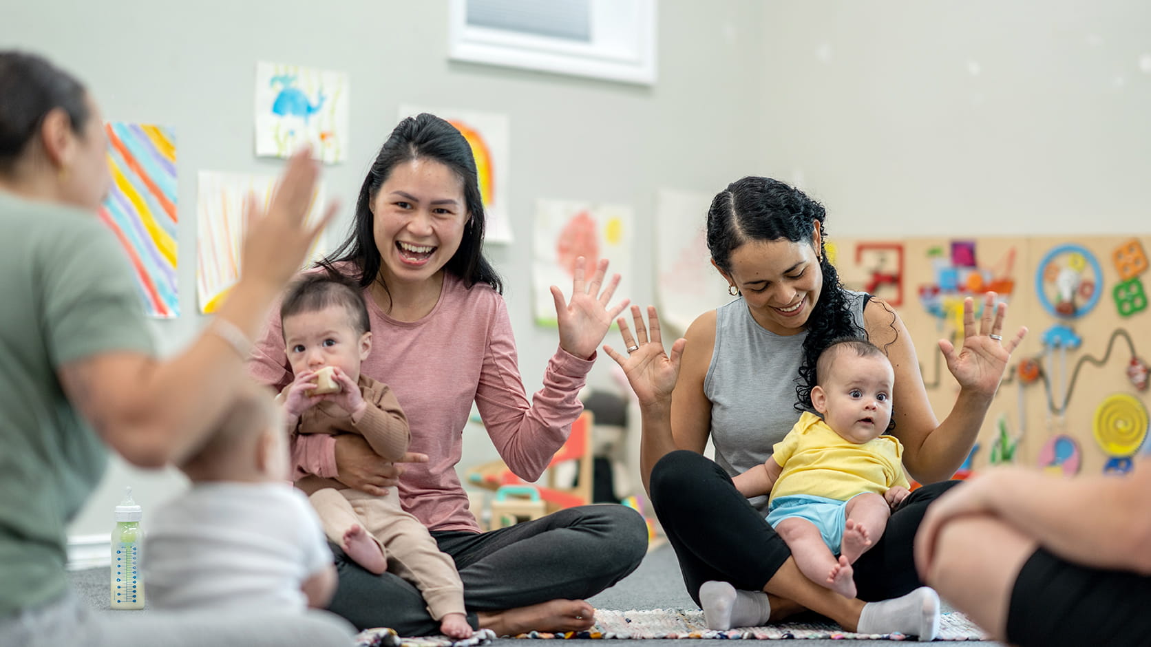 Mothers sit in a circle, smiling and waving their hands, while their babies sit in their laps.