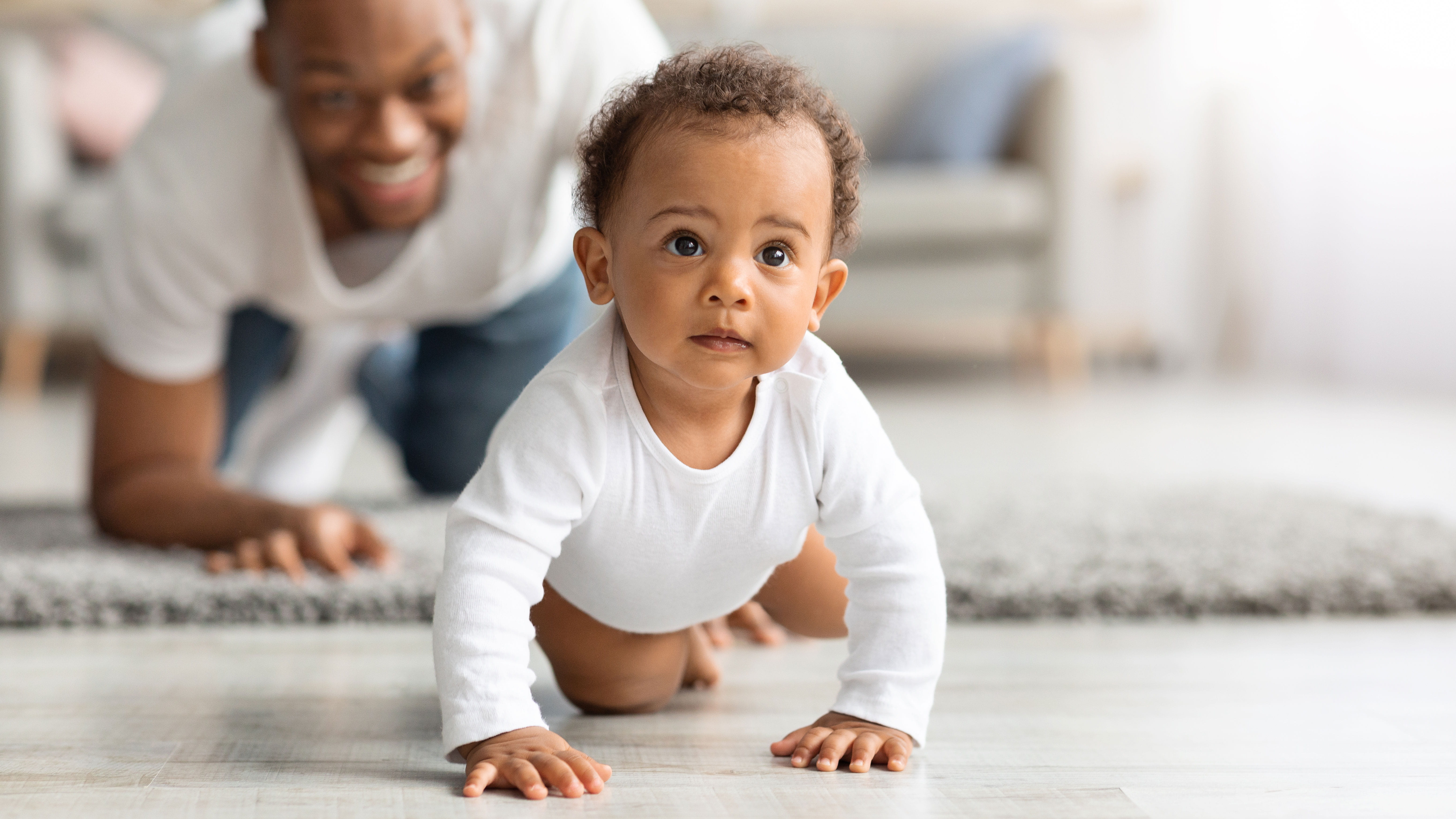 A baby crawls toward the camera while her dad smiles in the background.