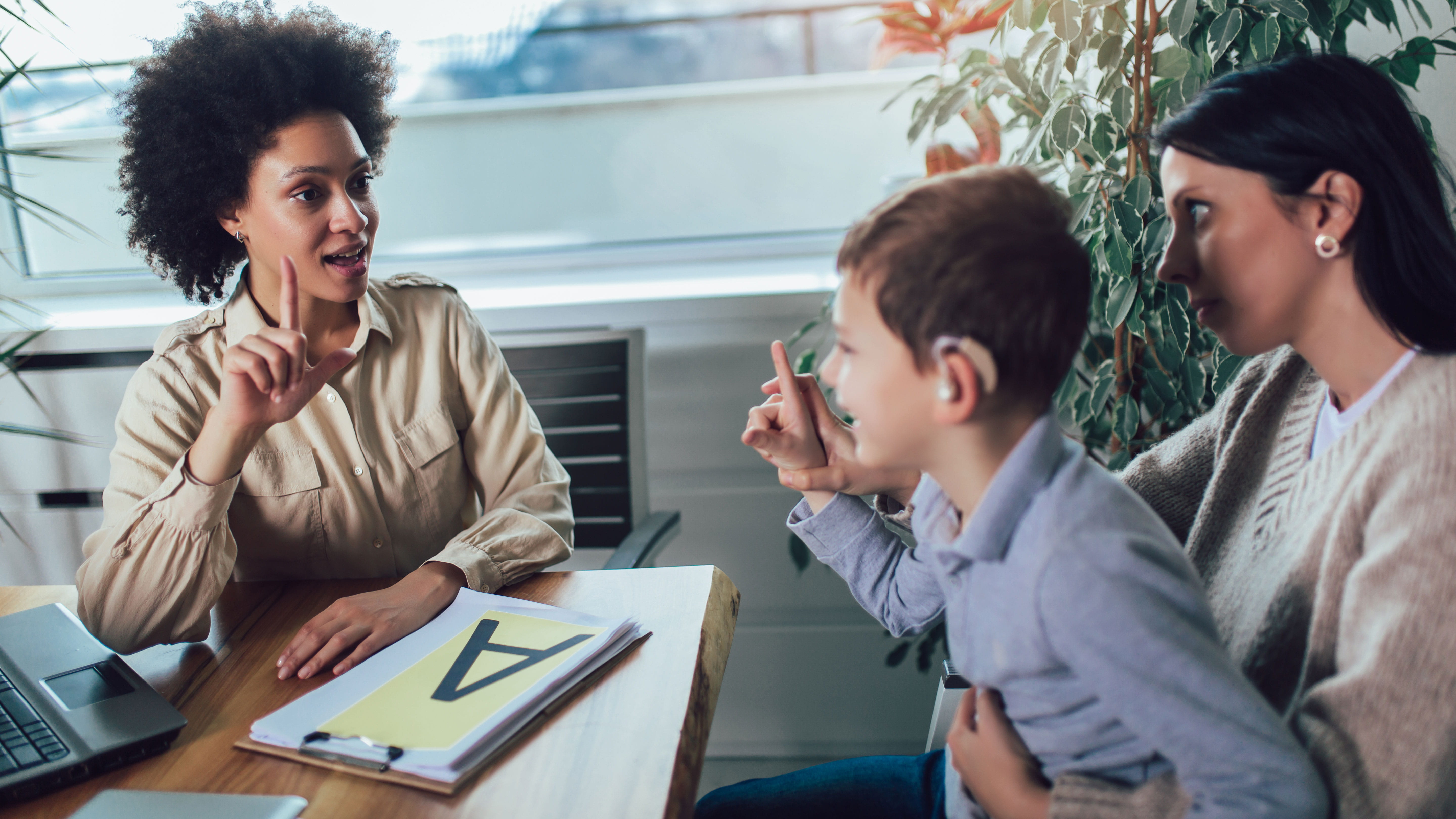 A teacher signs with a smiling young boy, who is wearing a hearing device and sitting in his mother's lap.