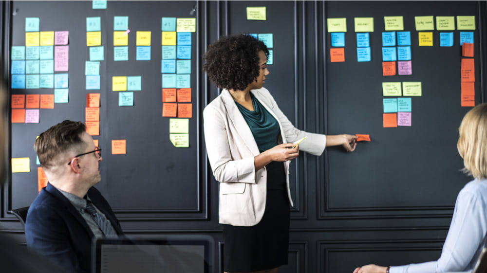 African American woman leading a meeting and placing sticky notes on a board.