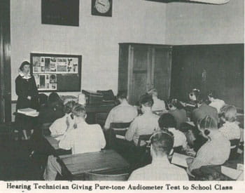 Black and white photo of of a hearing technician giving pure-tone audiometer test to a school class.