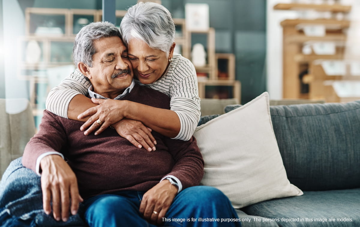Older woman standing behind an older man seated on a couch and hugging him from behind
