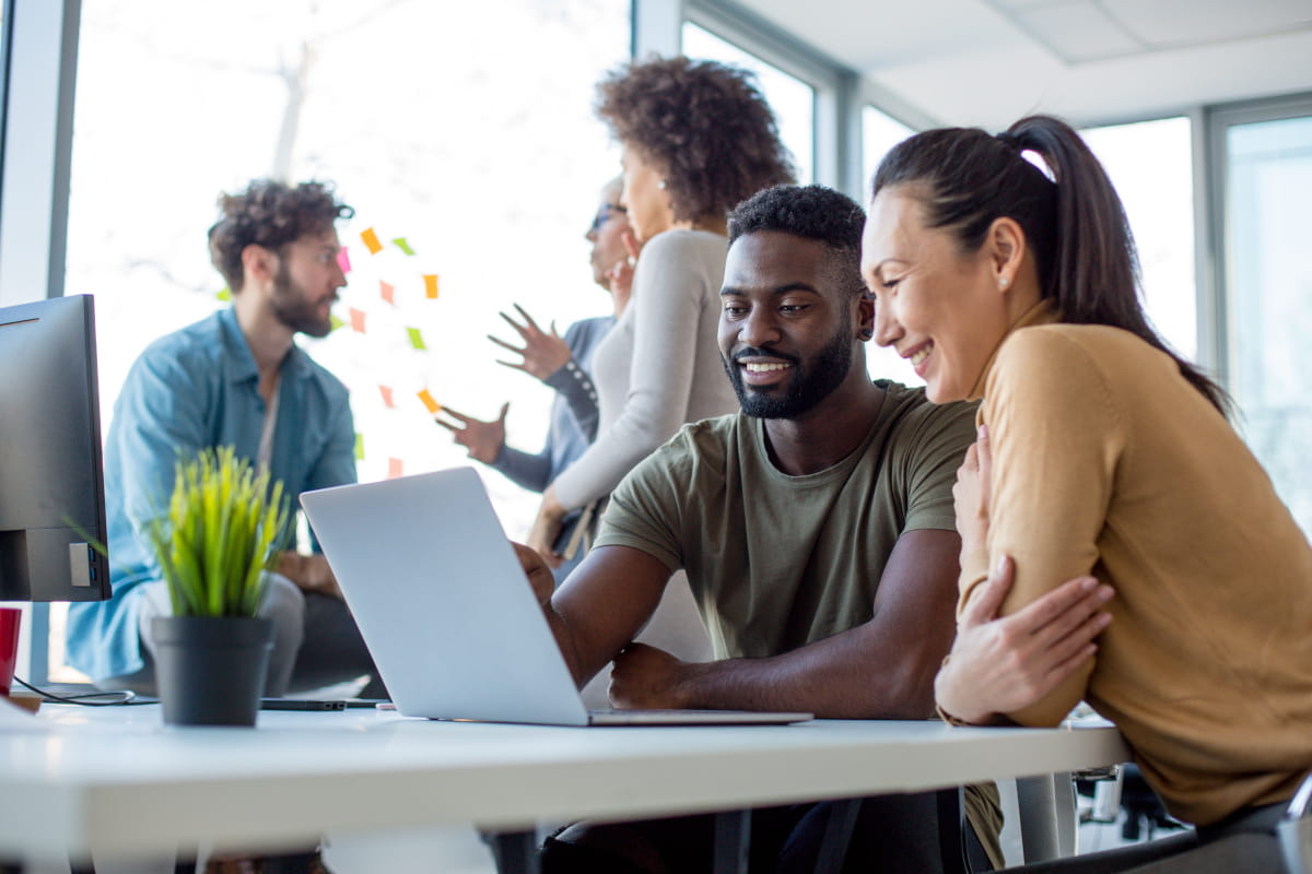 Multi-racial and multi-ethnic group of young people gathered in workplace, some standing by window, some seated in front of a laptop