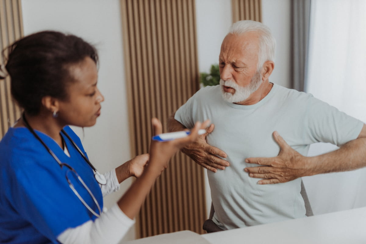 older man with white, short-cut beard seated on examination table, talking with female medical professional and gesturing to his chest