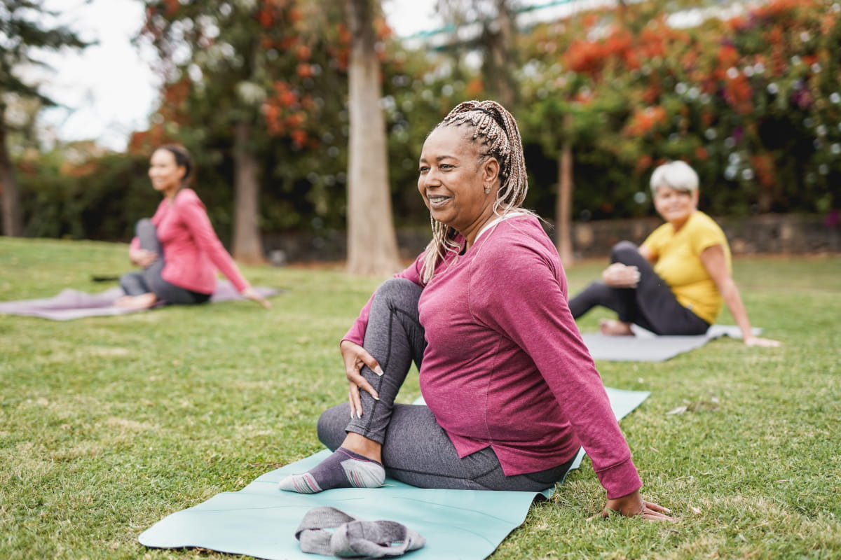 group of three middle-aged and older women seated on exercise mats in a grassy area of a park during exercise class