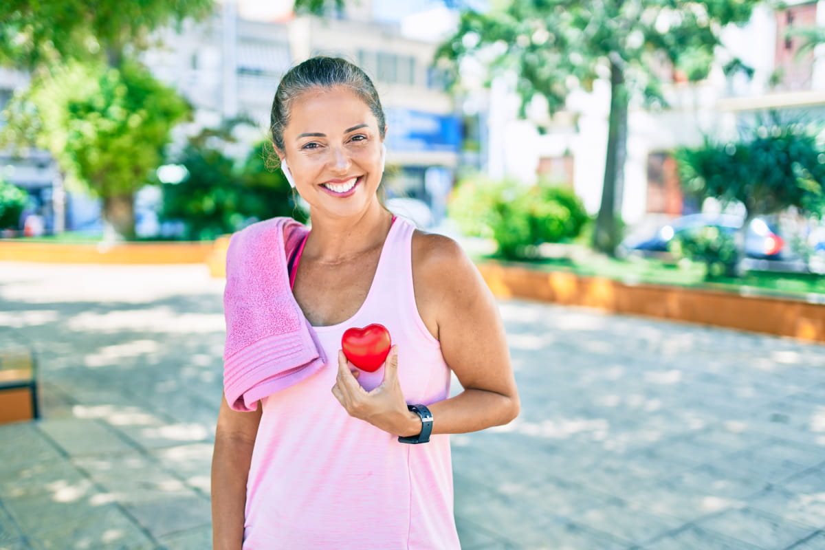 Middle-aged woman in park, wearing work-out clothing with towel over shoulder and replica of a heart in her hand