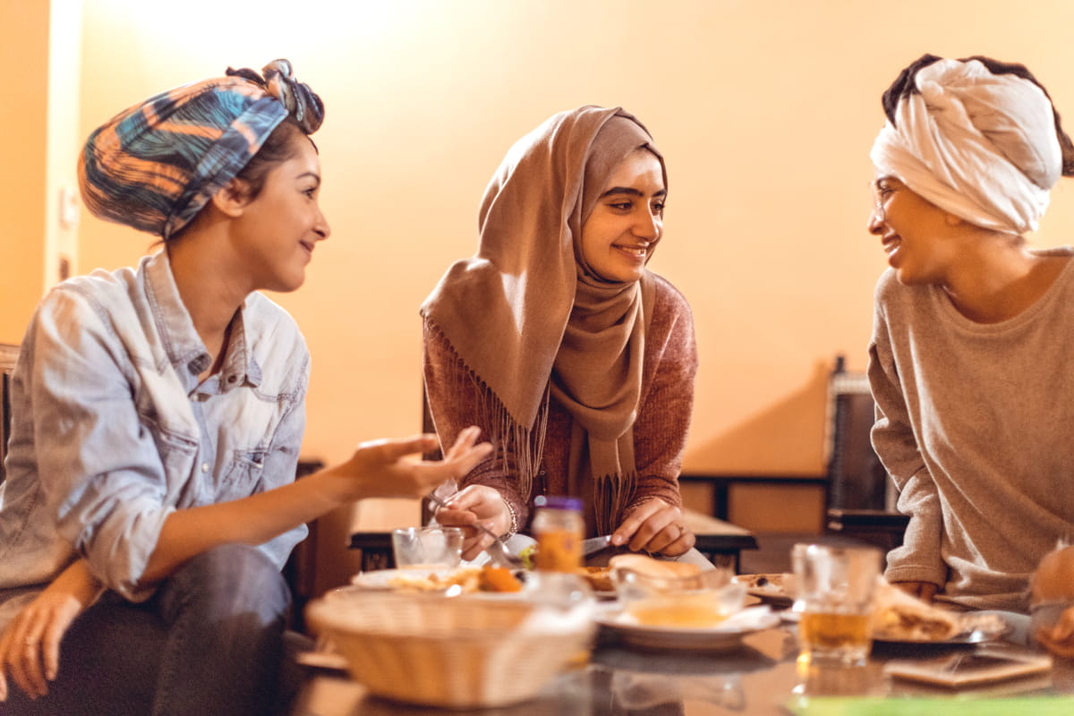 Three young women, wearing cloth head coverings, sitting at a table, eating and talking