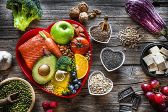 red heart-shaped dish holding a selection of healthy, portion-sized vegetables, grains and oils and surrounded by other vegetables, grains, and healthy foods