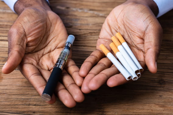 Man's hand holding vape and tobacco cigarettes over wooden desk