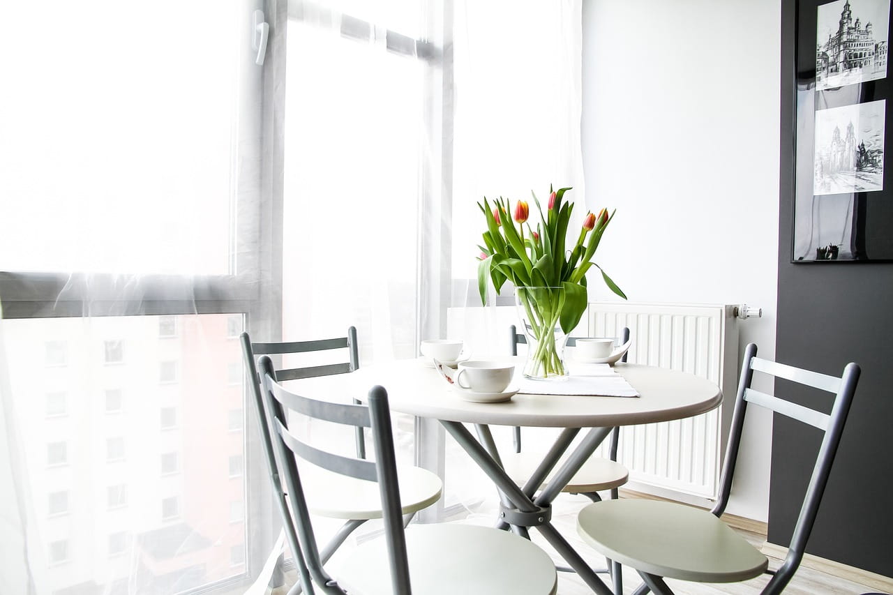 round white table with four white and gray chairs, three coffee cups and vase of tulips on table, sun coming through sheer-curtained window