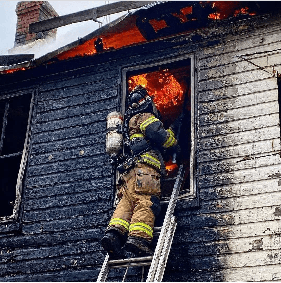 Firefighter on ladder outside upper story window with flames visible inside house, outside of house is scorched and blackened 