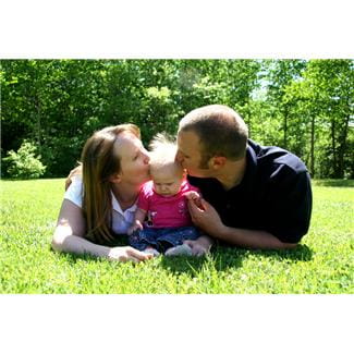 young white woman and man lying on their stomachs on the grass in the sunshine, kissing blonde-haired baby who is sitting between them