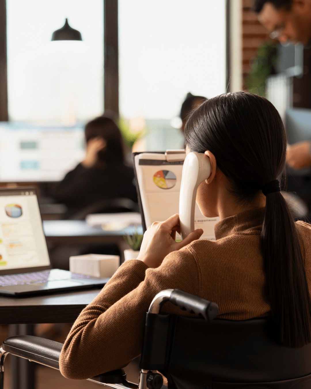 A workplace scene showing a woman using a captioned telephone while coworkers sit blurred in the background.