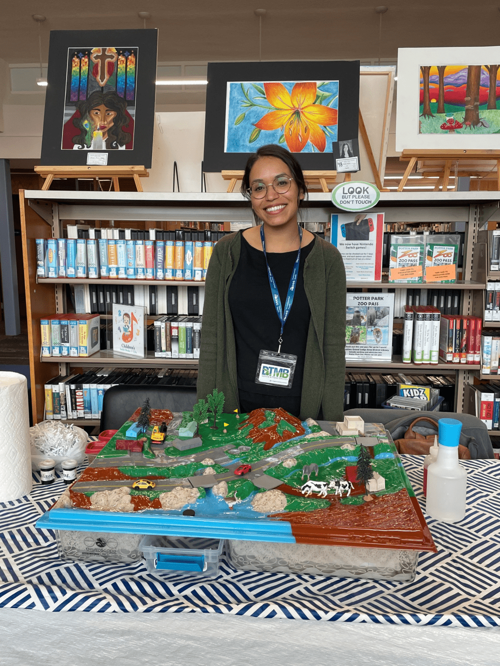 Woman standing behind a large model of a landscape with plastic houses, a factory, a farm, cars and roads, and a lake