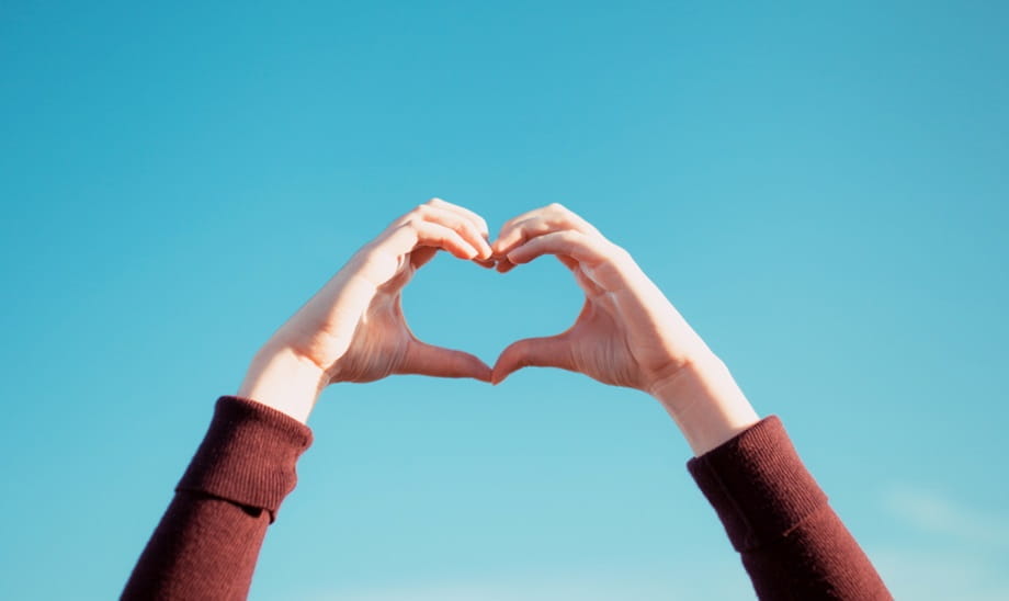 Hands being held up in a heart shape in front of a blue sky background