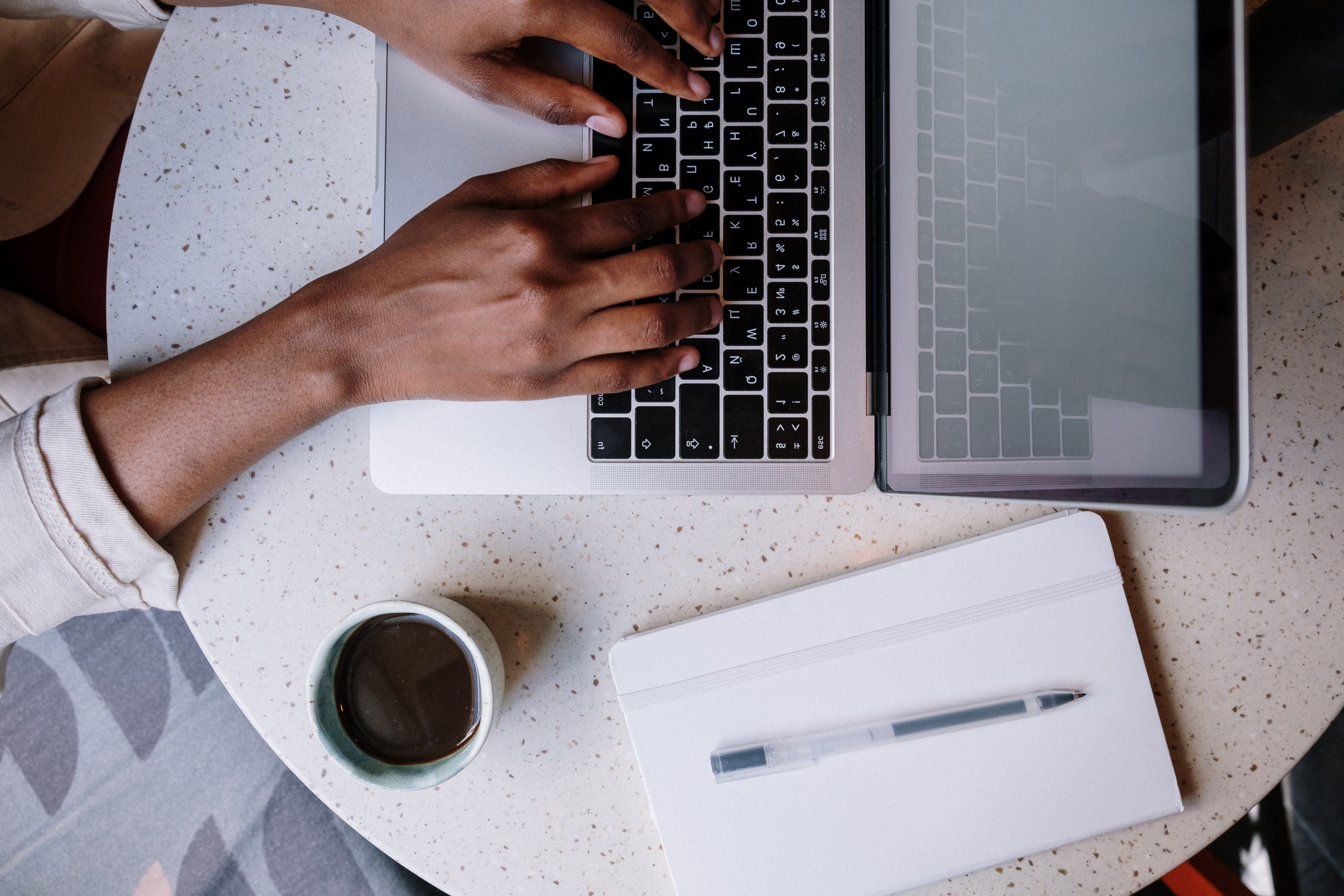 Overhead shot of s desk with a laptop that someone is typing on, a cup of coffee, and a notepad with pen.