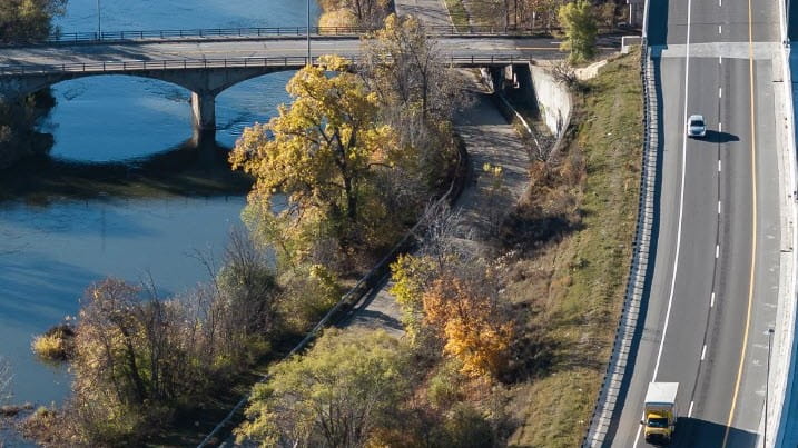 Aerial view of a river with a bridge crossing over it, surrounded by trees with autumn foliage. Next to the river is a curved pathway, and on the right side of the image a highway runs parallel, with a few vehicles traveling on it.