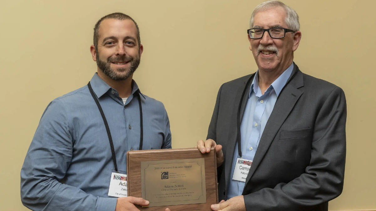 Two people stand side by side indoors, smiling and holding a wooden award plaque together. Both are wearing name badges and business attire against a plain beige background.