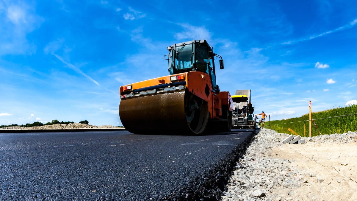 A close-up view of a road construction scene showing a large orange steamroller smoothing freshly laid asphalt under a bright blue sky. Construction workers and additional paving equipment are visible in the background along the roadside.