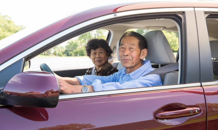 An older seat belted male driver with a female passenger sitting in a red vehicle.